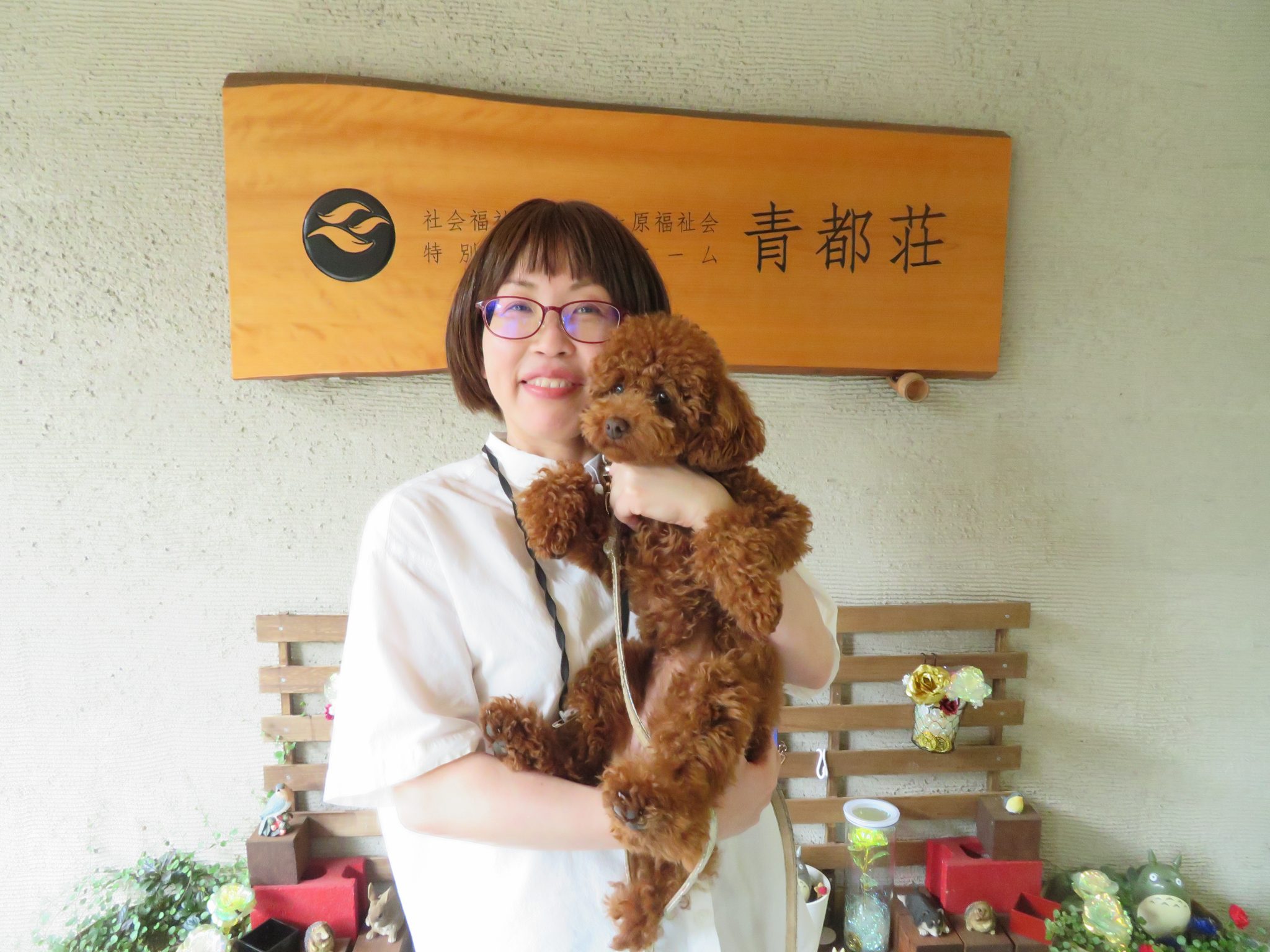 職員が施設内で小型犬を抱いて撮影。青都荘の看板が背景に見える。