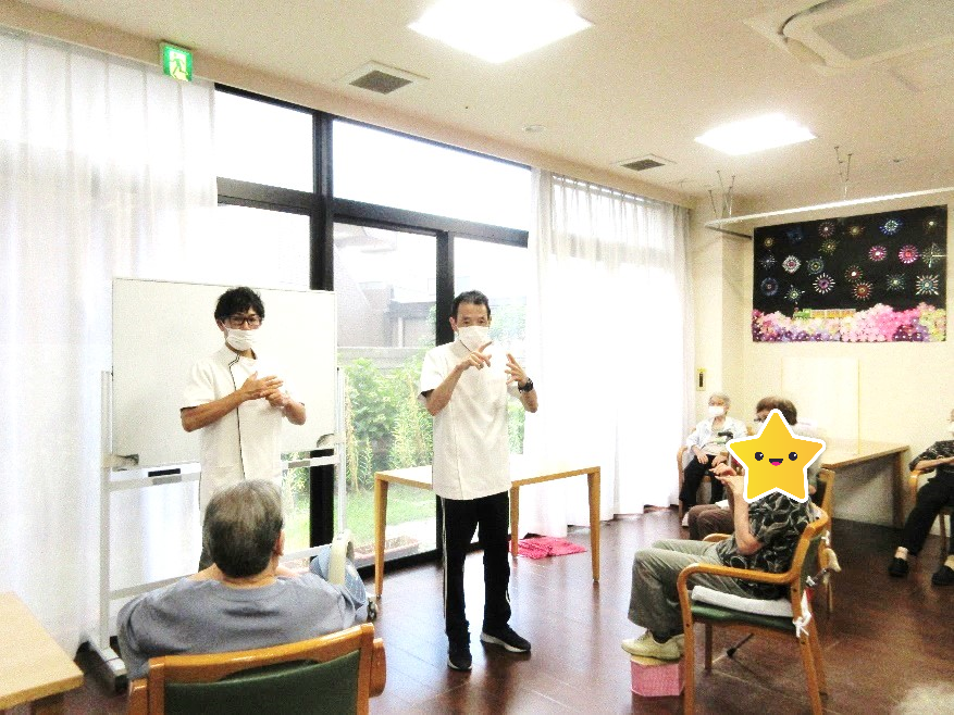 職員によるレクリエーション活動。入居者が見守る中、スタッフが手話のような動きで何かを演じている。